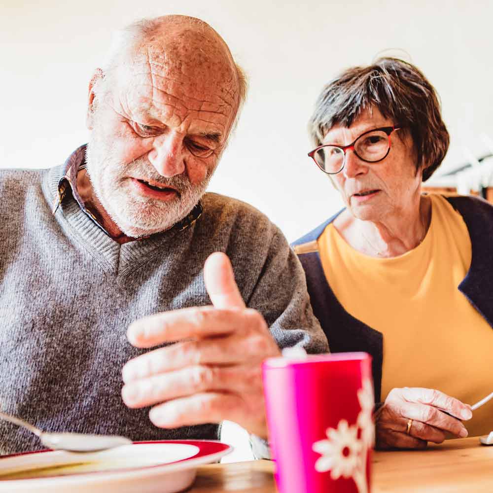 Ornamin-2CARE-Parkinson Älterer Mann und ältere Frau sitzen am Tisch nebeneinander
