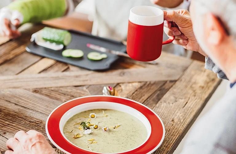 ORNAMIN-Demenzgeschirr-dementia tableware Auf dem Tisch steht ein Teller mit Kipp-Trick mit rotem Rand. On the table is a plate with special Features with a red rim.